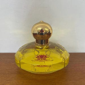 Könnte beinhalten: Eine klare, gelbe Parfümflasche mit einem goldfarbenen, kugelförmigen Verschluss. Die Flasche hat eine abgerundete, abgeflachte Form und ein rotes Blumendesign. Die Flasche steht auf einer Holzoberfläche.