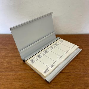 Könnte beinhalten: Ein grauer Metall-Schreibtischkalender mit Klappdeckel, geöffnet, um einen Wochenplaner zu zeigen. Die Planerseiten sind weiß mit schwarzen Linien und Text in mehreren Sprachen. Der Kalender liegt auf einer Holzoberfläche.