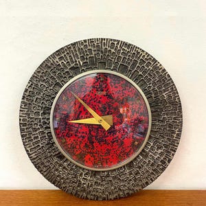 Könnte beinhalten: Eine runde Wanduhr mit einem texturierten, dunkelgrauen Rahmen in einem sich wiederholenden gitterartigen Muster. Das Zifferblatt ist leuchtend rot mit schwarzen Sprenkeln und durch eine Glasscheibe geschützt. Sie hat zwei spitze, goldfarbene Zeiger.