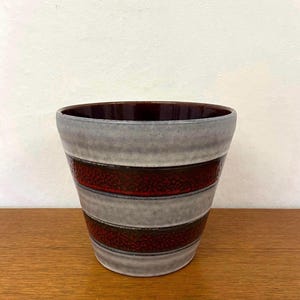 Pode incluir: Um vaso de cerâmica com formato cilíndrico cônico, apresentando faixas horizontais alternadas de vermelho escuro texturizado e esmalte cinza claro liso. O interior do recipiente possui um acabamento em vermelho escuro profundo e brilhante.