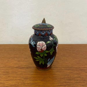 Pode incluir: Um pequeno vaso cloisonné preto com um desenho floral e uma tampa pontiaguda. O vaso é decorado com esmalte rosa, azul e verde.