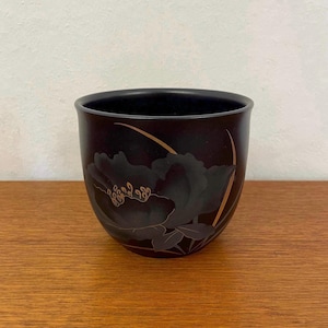 Può includere: Una tazza in ceramica nera con un disegno floreale dorato. La tazza è appoggiata su una superficie di legno.