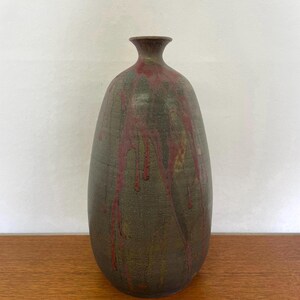 Könnte beinhalten: Eine hohe, ovale Keramikvase mit schmalem Hals. Die Vase hat eine dunkelgrüne Grundfarbe mit roten Tropfen, die die Seiten hinunterlaufen. Die Vase steht auf einer Holzoberfläche.