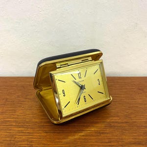Könnte beinhalten: Ein rechteckiger Europa-Reisewecker mit goldfarbenem Zifferblatt und schwarzen Zeigern. Die Uhr befindet sich in einem dunkel gefärbten, aufklappbaren Gehäuse, das ein metallisch goldenes Inneres enthüllt. Das Zifferblatt zeigt schwarze Ziffern.