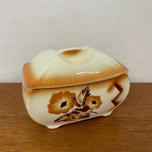 May include: Cream-colored ceramic trinket box with a lid. The box features a floral design in shades of brown and orange. The lid has a small handle and a decorative orange trim. The box is likely vintage.