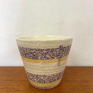 Pode incluir: Um vaso de cerâmica com uma superfície texturizada e salpicada, apresentando faixas horizontais em castanho, amarelo e verde pálido. O recipiente tem uma abertura larga e uma base cónica, com um acabamento rugoso e granular no exterior.