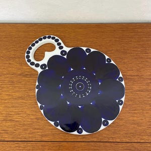 Op de afbeelding: Een ronde, witte keramische serveerschaal met een handvat, met een donkerblauw bloemmotief. Het handvat is versierd met kleine, gelijkmatig verdeelde blauwe stippen. Het ontwerp van de schaal omvat een centrale cirkel omgeven door bloembladachtige vormen.