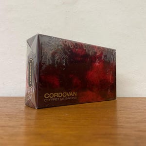 Op de afbeelding: Een rode en gouden doos met de tekst "Cordovan Coffret de Savons" op de voorkant. De doos is verpakt in plastic.
