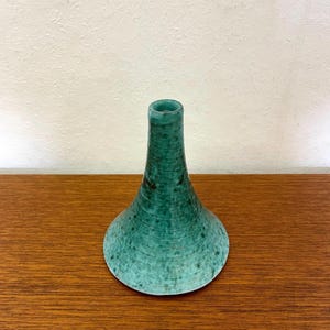 Op de afbeelding: Een blauwgroene keramische vaas met een brede, uitlopende voet die taps toeloopt in een smalle, cilindrische hals. Het oppervlak is voorzien van een gespikkeld glazuur met horizontale ribbeltexturen over het gehele lichaam van de vaas.
