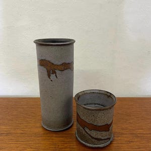 May include: A tall cylindrical ceramic vase and a shorter matching cylindrical ceramic cup. Both pieces feature a textured grey finish with a distinct, horizontal band of metallic copper-toned glaze that appears to drip down the sides.
