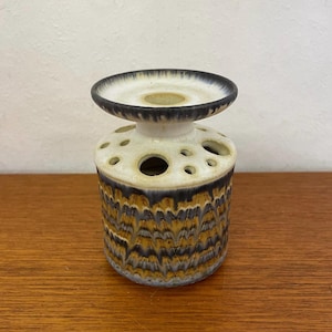 May include: A white ceramic bud vase with a brown and blue glaze pattern. The vase has a round base with a ring of holes around the top and a small, flat, circular top.