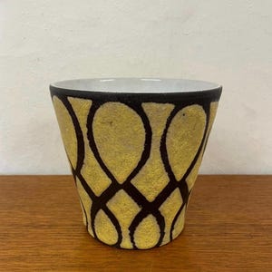 Könnte beinhalten: Ein Keramik-Pflanzgefäß mit einem gelb-schwarzen geometrischen Muster. Der Pflanztopf hat einen weißen Innenraum und einen schwarzen Rand. Das Design zeigt miteinander verbundene Tropfenformen in Gelb, umrandet von schwarzen Linien. Der Pflanztopf steht auf einer Holzoberfläche.