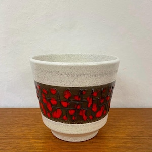 Pode incluir: Um vaso de cerâmica com uma seção superior branca salpicada e uma faixa marrom com formas vermelhas irregulares. O vaso tem uma borda ligeiramente alargada e uma base de pedestal.