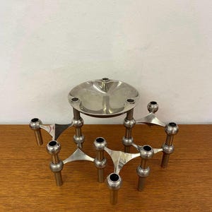 Può includere: Un portacandele modulare in metallo color argento con molteplici supporti circolari interconnessi e un vassoio circolare centrale rialzato. La struttura è composta da diversi segmenti collegati tra loro a forma di treppiede.
