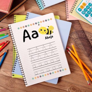 May include: A spiral-bound notebook with the letter "A" and the word "Abeja" with a cartoon bee illustration. The notebook has lines for practicing writing the letter "A". Pencils, a calculator, and other notebooks are in the background.