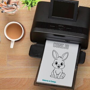 May include: A black printer is printing a coloring page with a cartoon bunny and the word "CONEJO". A cup of coffee sits on a wooden coaster next to the printer. Pencils and a small plant are also visible.