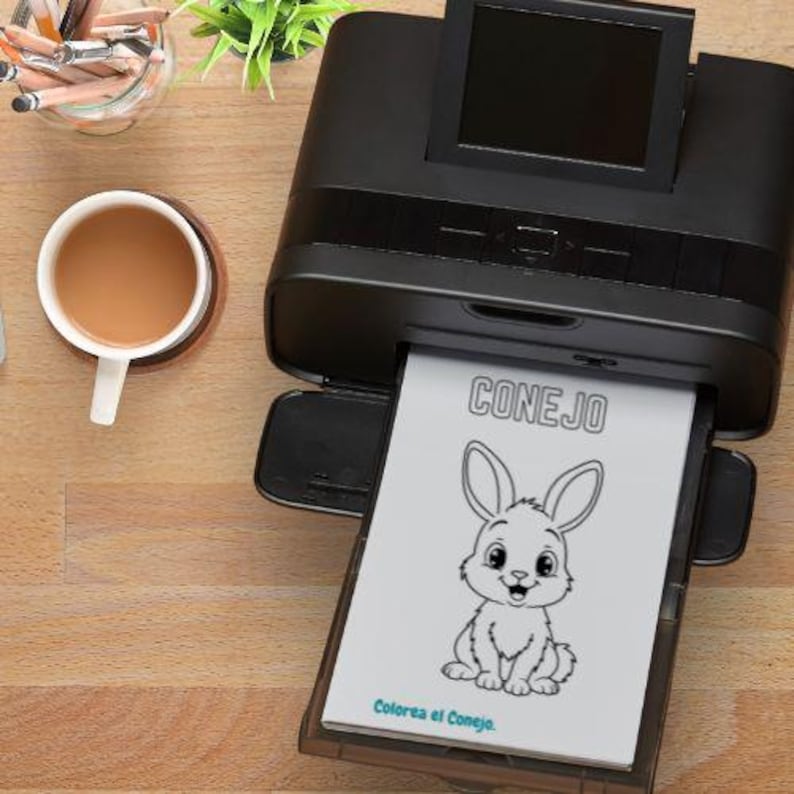 May include: A black printer is printing a coloring page with a cartoon bunny and the word "CONEJO". A cup of coffee sits on a wooden coaster next to the printer. Pencils and a small plant are also visible.