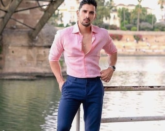 May include: A man wearing a pink linen shirt, dark blue pants, and white sneakers. He is standing by a railing with a body of water behind him.