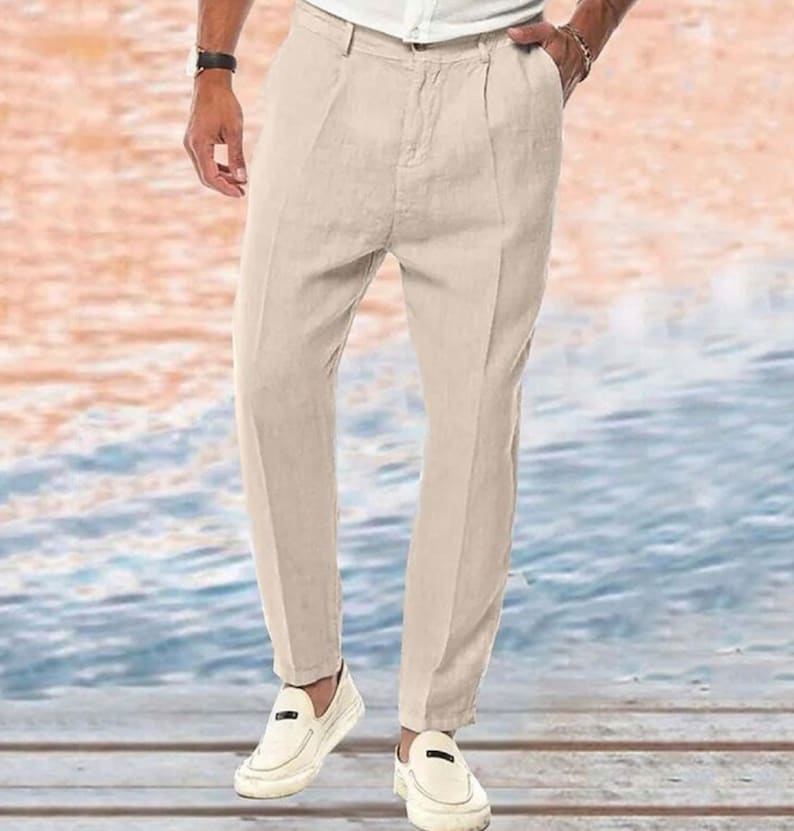May include: A pair of light beige linen pants with a straight leg and a slightly cropped length. The pants have a front pleat detail and a zipper fly closure.