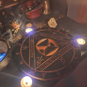 K&ouml;nnte beinhalten: Ein schwarzer Holzkreis mit einem geometrischen Design und den Worten "ERESHKIGAL", "AKTOPHIS" und "NEBOUTOSQALETH", die um den Rand herum eingeschrieben sind. Der Kreis ist von vier brennenden Kerzen umgeben.