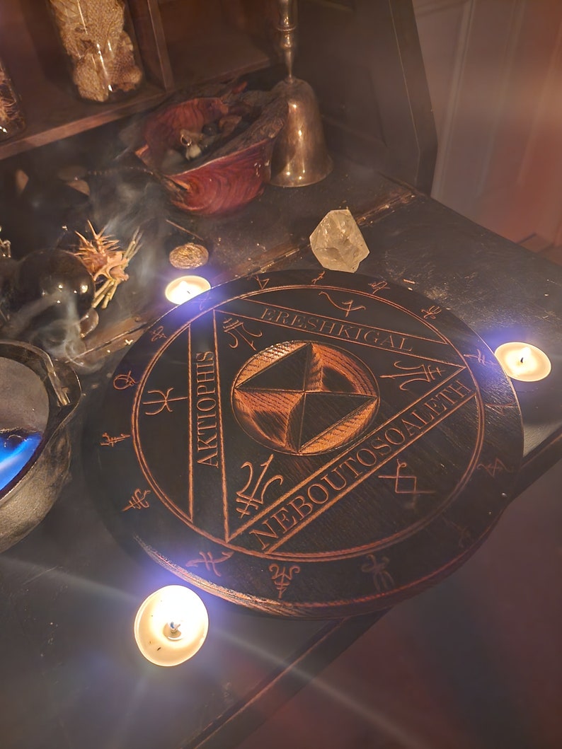 K&ouml;nnte beinhalten: Ein schwarzer Holzkreis mit einem geometrischen Design und den Worten "ERESHKIGAL", "AKTOPHIS" und "NEBOUTOSQALETH", die um den Rand herum eingeschrieben sind. Der Kreis ist von vier brennenden Kerzen umgeben.