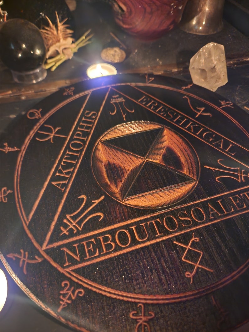 K&ouml;nnte beinhalten: Ein schwarzer Holzkreis mit einem geschnitzten Design, das ein Dreieck mit den W&ouml;rtern "Aktophis", "Neboutoscale" und "Ereshkigal" zeigt, die um ihn herum eingeschrieben sind. Der Kreis hat eine dunkle Holzmaserung und eine r&ouml;tlich-braune F&auml;rbung.