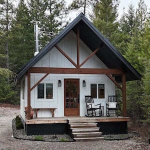 Może przedstawiać: Mała, biała chatka z czarnym dachem i drewnianymi akcentami. Chata ma werandę z dwoma krzesłami bujanymi i małą ławką. Drzwi wejściowe są brązowe, a po obu stronach znajdują się okna. Chata jest otoczona drzewami.