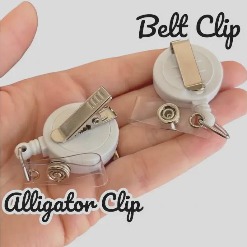 May include: Two white retractable badge reels featuring a circular housing. One reel has a metal alligator clip, and the other has a metal belt clip. Both include a clear plastic strap with a metal snap button for attaching identification cards.