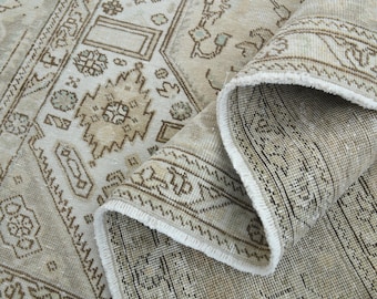 May include: Beige and brown vintage-style rug with intricate geometric patterns and a rolled-up edge. The rug features a distressed look with a mix of light and dark brown tones, creating a textured appearance.