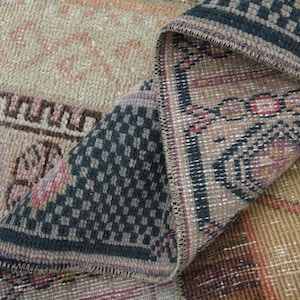 May include: A close-up of a folded rug with a geometric pattern in shades of brown, black, and pink. The rug has a fringe border and is made of woven fabric.