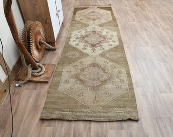 May include: A rectangular, olive green runner rug with a geometric pattern in beige and pink. The rug features a central design of diamonds and octagons. The rug is placed on a wooden floor.