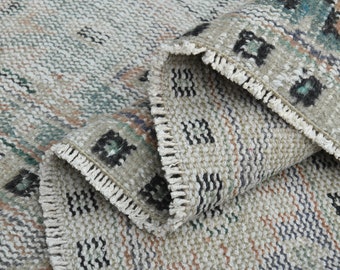 May include: A close-up of a vintage rug with a folded corner, showcasing a textured weave. The rug features a neutral color palette with accents of teal, orange, and black geometric patterns. The edges have a fringed trim.