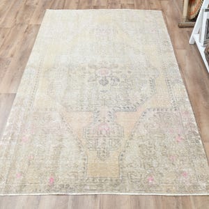 Puede incluir: Una alfombra rectangular de estilo vintage con un aspecto descolorido y desgastado. La alfombra presenta un diseño central de medallón en tonos apagados de beige, rosa y gris. Está colocada sobre un suelo de madera.