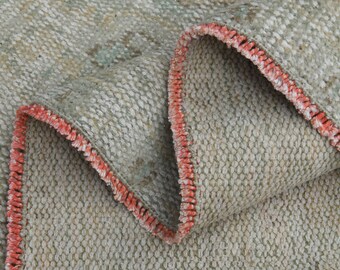 May include: Close-up of a light gray woven rug with a red fringe border. The rug is folded in half, showing the texture of the fabric.