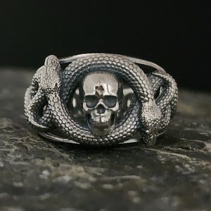 May include: A silver ring featuring a detailed skull design with a snake wrapped around it.