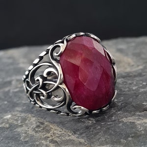 May include: A silver ring with an ornate design featuring a large, oval-shaped, faceted, red gemstone.