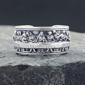 Può includere: Un anello d'argento con una rappresentazione dettagliata dell'Ultima Cena. L'anello è largo e presenta un design in rilievo di Gesù e dei suoi discepoli attorno a un tavolo.