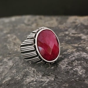 May include: A silver ring with a large, oval-shaped, red gemstone. The ring has a textured band with vertical lines.