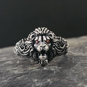 May include: A silver ring with a detailed lion head design. The lion has red eyes and an open mouth. The ring is on a gray textured surface.