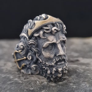 Peut inclure: Une bague en argent avec une sculpture détaillée d'un visage d'homme avec une barbe. L'homme porte une couronne avec un motif de trident doré. La bague est posée sur une surface en pierre grise.