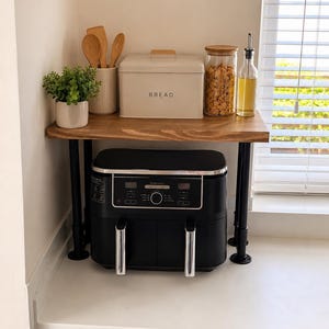 May include: A black dual-basket air fryer sits under a wooden shelf supported by black metal pipes. On the shelf, there is a cream-coloured metal bread box with the word BREAD printed on it, a glass jar with cereal, a glass oil bottle, and a plant.
