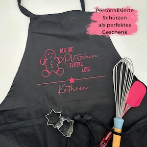 Könnte beinhalten: Schwarze Schürze mit einem rosafarbenen Lebkuchenmann-Motiv und dem Text "Auf die Plätzchen fertig, los!" und "Kathrin".