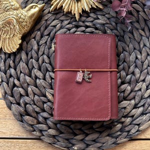 Op de afbeelding: Een bordeauxrood leren notitieboek met een bruine elastische sluiting en decoratieve bedels. Het notitieboek is gesloten en heeft een gestikte rand. Een kleine bedel van een vogel en een envelop hangt aan het elastiek. Het notitieboek ligt op een geweven mat.
