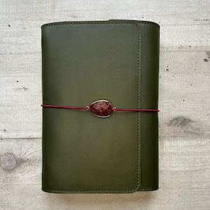 Può includere: Una copertina rettangolare per taccuino in pelle verde oliva con finitura liscia e opaca e bordi cuciti. Un sottile cordino elastico rosso scuro avvolge orizzontalmente il centro, fissato da una singola gemma rosso scuro sfaccettata a goccia.