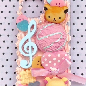 Könnte beinhalten: Eine transparente Handyhülle, verziert mit verschiedenen pastellfarbenen Charms. Das Design umfasst einen hellblauen Notenschlüssel, eine rosa Eiskugel, ein Herz mit weißen Punkten und bärenförmige Figuren. Der Hintergrund ist ein weißes und schwarzes Tupfenmuster.
