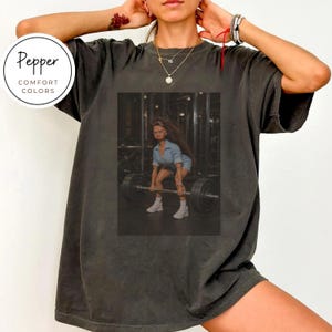 Könnte beinhalten: Ein anthrazitfarbenes Kurzarm-T-Shirt mit einem rechteckigen Grafikdruck einer Puppe in blauem Jeans-Outfit, die eine schwere Langhantel in einem Fitnessstudio hebt. Das T-Shirt hat einen Rundhalsausschnitt und eine lockere, entspannte Passform.