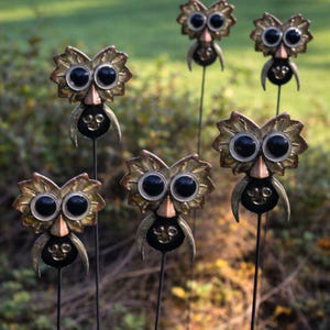 May include: Six metal owl garden stakes are displayed. Each stake features an owl with large black eyes, a copper-coloured beak, and a smiling face. The owl's head has a leaf-like design with gold and bronze tones.