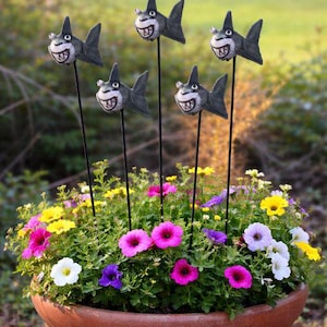 May include: A terracotta plant pot filled with vibrant flowers in shades of pink, yellow, white, and purple. Five novelty shark garden ornaments with grey bodies and smiling faces are placed amongst the flowers.