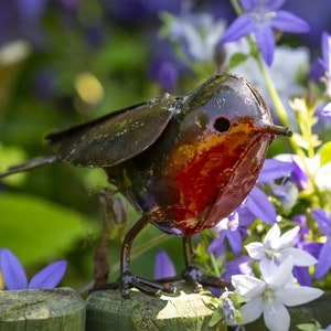 May include: A metal garden sculpture of a robin perched on a wooden fence post. The robin is painted in shades of brown, black, and red, with a realistic design. The sculpture is surrounded by purple and white flowers.