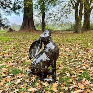 May include: A black metal sculpture of a rabbit with its ears up, sitting on a bed of fallen leaves.